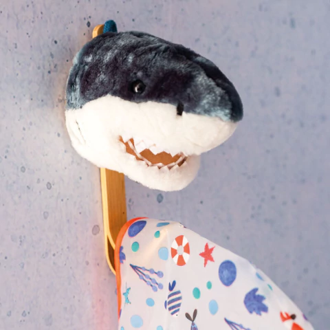 Cabide de parede infantil em formato de cabeça de tubarão de pelúcia Wild & Soft, decorativo, pendurado em parede azul, com roupa colorida pendurada.