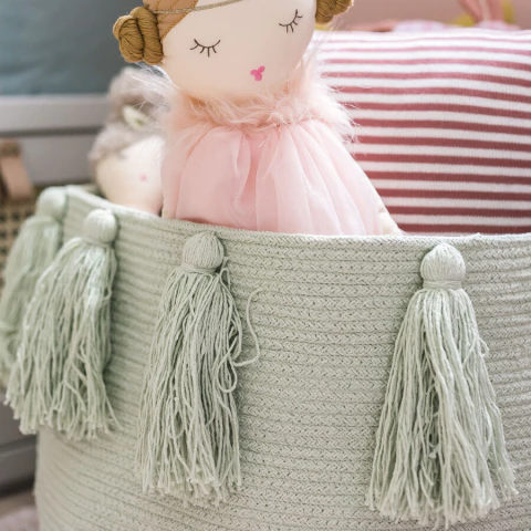 Cesto lavável verde com franjas decorativas da Lorena Canals, usado para organização e decoração, com boneca de pano dentro.