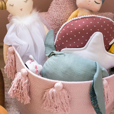 Cesto lavável rosa com franjas da Lorena Canals, decorado com almofadas e brinquedos infantis em tons suaves.