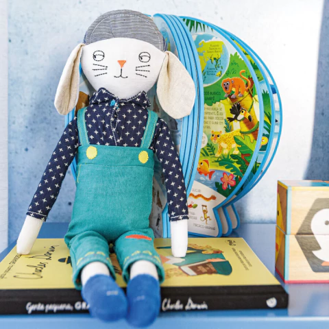 Boneco de pelúcia em formato de coelho com jardineira azul, camisa de estrelas e gorro, sentado sobre livros infantis em um ambiente decorativo.