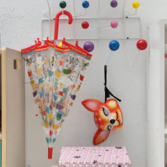 Guarda-chuva infantil colorido com estampa de crianças e animais, da marca Djeco, pendurado em cabideiro ao lado de uma máscara de cervo em ambiente infantil.