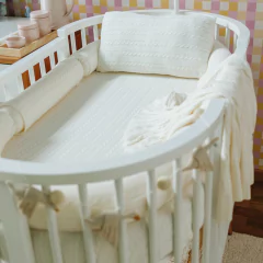 Kit berço Rian Tricot 5 peças na cor off-white, com proteção lateral, travesseiro e manta, decorando um berço oval branco em ambiente infantil.