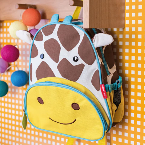 Mochila infantil Skip Hop Zoo modelo girafa, com detalhes amarelos, marrom e branco, pendurada em cabideiro escolar.
