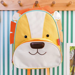 Mochila infantil Zoo Leão da Skip Hop, com design divertido de leão em tons de amarelo e laranja, pendurada em cabideiro.