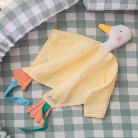 Naninha de algodão natural em formato de pato, com corpo amarelo claro e detalhes coloridos, sobre superfície xadrez verde e branca. Brinquedo seguro e macio para bebês.