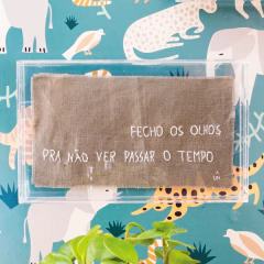 Quadro decorativo com caixa de acrílico e linho terroso, exibindo a frase "Fecho os olhos pra não ver passar o tempo", posicionado em frente a uma parede com estampa de animais coloridos.