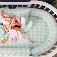 Rolo de berço em tricô vichy verde claro, usado como protetor de berço, com almofada e bonecas decorativas dentro de um berço branco.