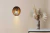 Aplique PRAGA en madera MDF, con diseño ovalado y tiras de madera, iluminando un ambiente decorado con un jarrón de flores secas.