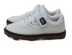 Tenis Klin brancos com fecho de velcro e solado marrom, posicionados sobre um fundo peludo.