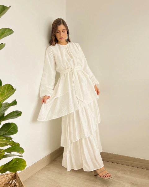 Vestido T.153 Blanco de PRIMIA, vista frontal mostrando la falda larga en capas, los volantes diagonales y la cintura definida con cinturón.