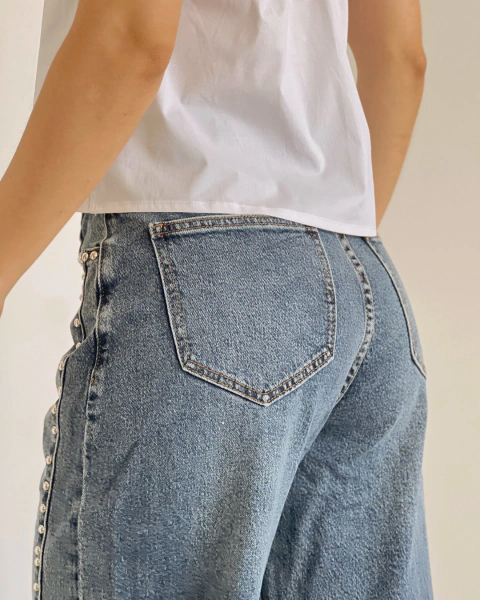 Jean azul de PRIMIA, primer plano posterior del bolsillo trasero con costuras visibles, lavado desgastado y aplicaciones metálicas en el lateral.