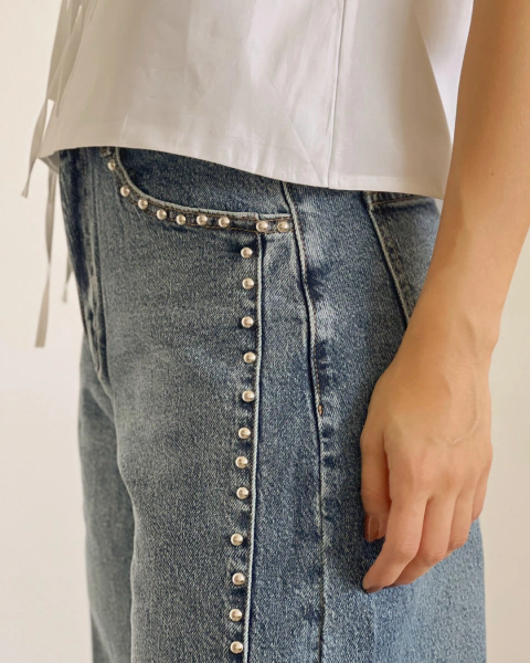 Jean azul de PRIMIA, primer plano de la cintura y el costado con detalles metálicos plateados distribuidos verticalmente y acabado denim desgastado.