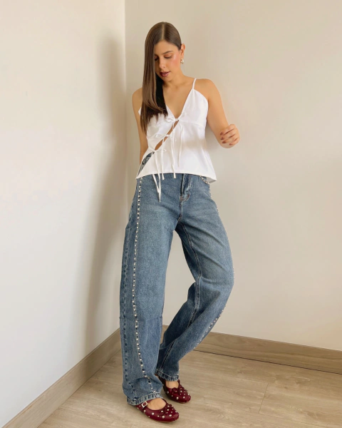 Jean azul de PRIMIA, vista frontal en ángulo con silueta de pierna ancha, acabado denim desgastado y aplicaciones metálicas decorativas en ambos laterales.