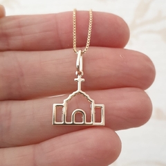 Colar folheado a ouro com pingente em formato de igreja vazada, segurado na mão.