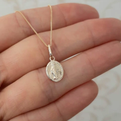 Colar folheado em ouro com medalha milagrosa, segurado por uma mão.