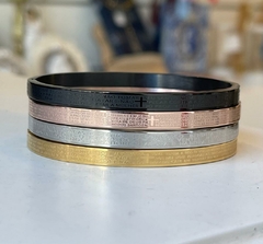 Braceletes de aço em quatro cores (preto, rose, prata, dourado) com gravações de orações, empilhados sobre a mesa.