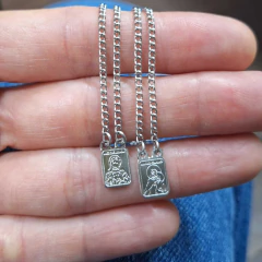 Escapulários retangulares pequenos de aço com pingentes dupla face, apresentados em corrente de 60 cm, exibidos sobre a palma da mão