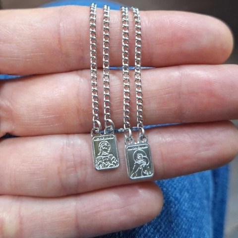 Escapulários retangulares pequenos de aço com pingentes dupla face, apresentados em corrente de 60 cm, exibidos sobre a palma da mão