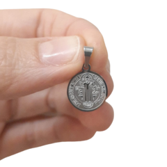 Pingente medalha de São Bento em aço, medindo 1,3 cm, segurado por uma mão.