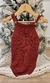Body vermelho de bebê com alças e botão, pendurado em um cabide, em frente a uma árvore de Natal decorada.