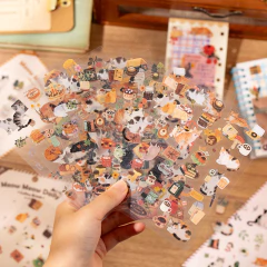 Mano sosteniendo planchas de stickers con ilustraciones de gatos y objetos cotidianos en tonos cálidos