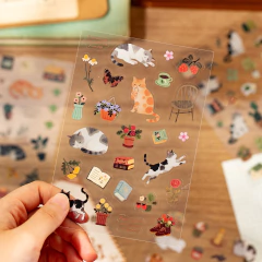 Hoja de stickers transparentes con ilustraciones de gatos, flores, libros y objetos decorativos sostenida por una mano sobre un fondo de escritorio de madera