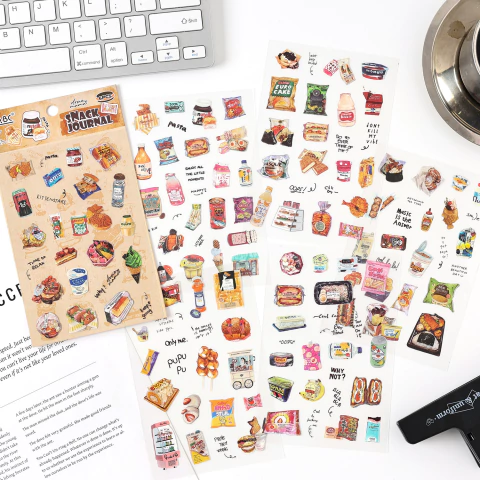 Pack de seis planchas de stickers con ilustraciones de snacks, alimentos y productos variados, sobre una mesa blanca junto a un teclado y una taza