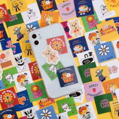 Un teléfono transparente con stickers coloridos sobre una superficie llena de stickers variados, incluyendo el texto "KEEP ON SMILING".