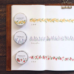 Rollos de washi tape troquelada con diseños florales y frutas sobre un cuaderno abierto, mostrando cintas decorativas de mimosa amarilla, lavanda morada y frutillas rojas.