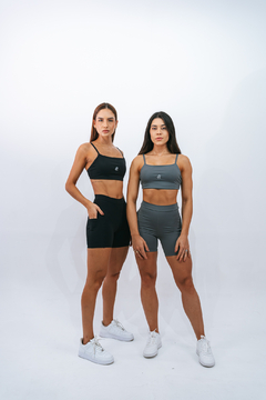 Duas mulheres usando shorts e tops esportivos, uma vestindo shorts pretos e a outra em tons de cinza, em um fundo branco.