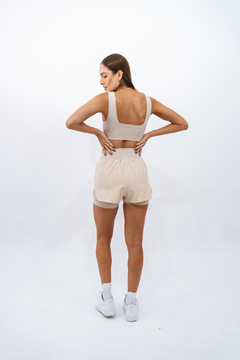 Modelo feminina usando shorts bege Duna Lurk, vista de costas, em ambiente neutro.