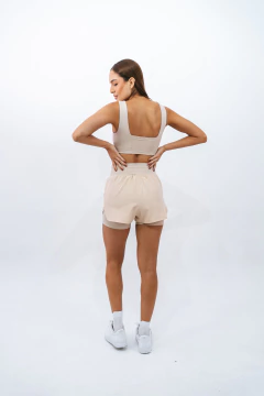 Modelo feminina usando top e shorts em tom bege, vista de costas, com fundo branco.