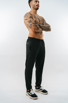 Modelo masculino usando calça tactel preta, com tatuagens visíveis e tênis esportivos.