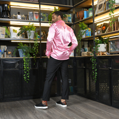 Hombre de espaldas vistiendo una camisa satinada rosa y pantalones negros, posando en un entorno moderno con estanterías llenas de plantas y objetos decorativos.