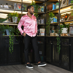 Hombre de pie, vistiendo una camisa satinada rosa y pantalones negros, en un ambiente moderno con plantas decorativas y estanterías.