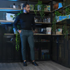Hombre de pie en una habitación moderna, vestido con un suéter negro y pantalones verdes, rodeado de plantas y estanterías con productos.