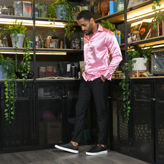 Hombre con camisa rosa y pantalones negros de pie en un entorno con plantas y estanterías decorativas.
