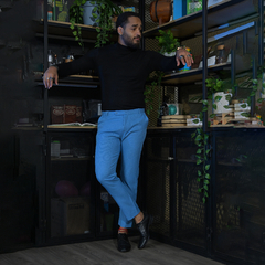 Hombre de pie con pantalones azules y camiseta negra, apoyado en un mueble con plantas y decoraciones.