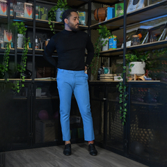 Hombre de pie en un entorno moderno, vistiendo un suéter negro y pantalones azul claro, rodeado de plantas y estantes con libros y objetos decorativos.