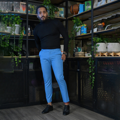 Hombre posando con un suéter negro y pantalones azul claro en un ambiente decorado con plantas y estanterías.