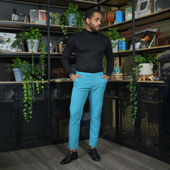 Hombre con pantalones lino azul turquesa y camiseta negra, de pie en un ambiente decorado con plantas y estantes.
