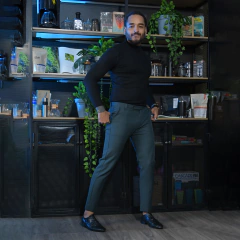 Hombre posando con un suéter negro y pantalones verdes frente a estanterías con plantas y accesorios de café.