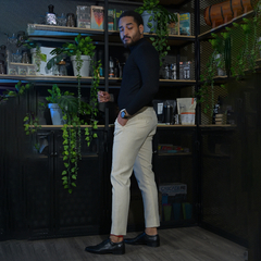 Hombre de pie en un entorno moderno, vistiendo un suéter negro y pantalones claros, con plantas decorativas y estanterías de fondo.