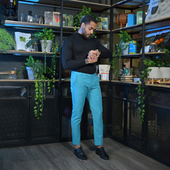 Hombre con camisa negra y pantalones lino azul claro, revisando su reloj en un ambiente moderno con estanterías y plantas.