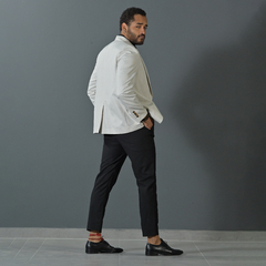 Hombre con blazer premium slim fit color tiza, de pie en un fondo gris, luciendo pantalones negros y zapatos elegantes.