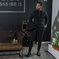 Hombre vestido con un traje negro, de pie junto a un sofá de cuero marrón y una mesa de madera, en un ambiente moderno.