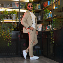Hombre con traje beige y gafas de sol, posando de pie en un entorno moderno con plantas y estanterías.