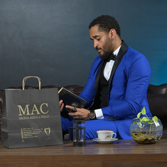 Hombre sentado en un sofá con traje azul, leyendo un libro, junto a una bolsa de compras de Moda Azul Cielo y una taza de café.