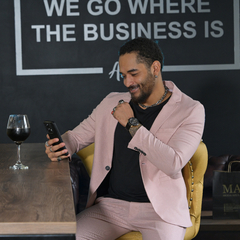 Hombre sentado en un bar con traje rosa, mirando su teléfono y sonriendo, con una copa de vino tinto en la mesa.