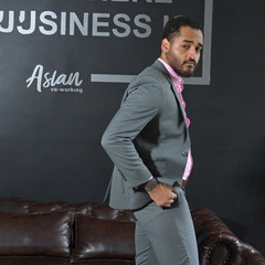 Hombre vestido con traje gris y camisa rosa, posando con una mano en el bolsillo, en un entorno de oficina moderna.